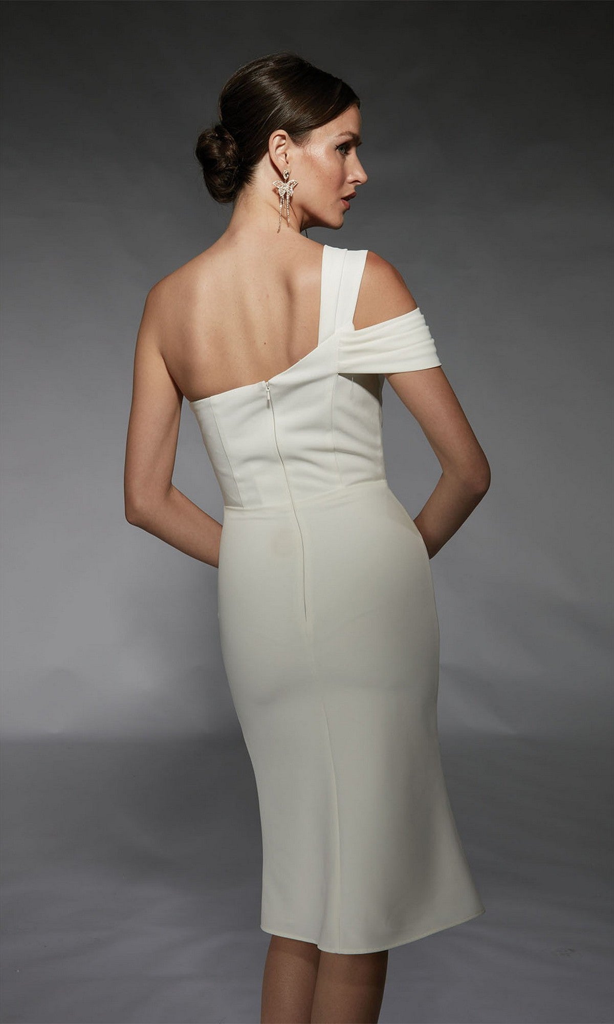 A woman in the Alyce 70054 knee-length semi-formal dress, featuring a fitted off-the-shoulder white design, stands with her back to the camera against a dark background.